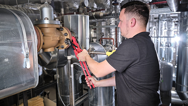 Ein Mann arbeitet mit einer Rohrzange an Heizungsanlage in einem industriellen Maschinenraum