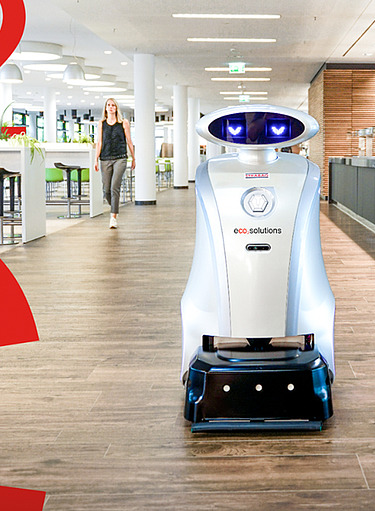 Autonomer Reinigungsroboter mit ‚eco solutions‘-Aufschrift in einem hellen, offenen Raum  mit großen Fenstern, Gemeinschaftsbereich mit Tischen und Theke, im Vordergrund rote CO2 Buchstaben