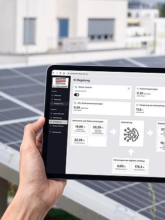 Obrázek ruky držící tablet s naměřenými hodnotami a stupnicemi, s fotovoltaickým systémem v pozadí