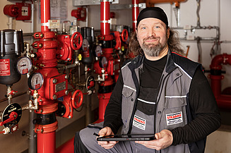 Implementierung STRABAG PFS Österreich Ein STRABAG-Techniker mit Tablet steht vor roten Rohrleitungen und Ventilen in einem technischen Raum..