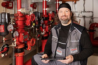 Ein STRABAG-Techniker mit Tablet steht vor roten Rohrleitungen und Ventilen in einem technischen Raum..