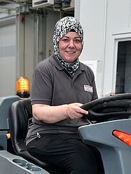 Frau mit Kopftuch fährt ein Reinigungsfahrzeug in industrieller Umgebung.