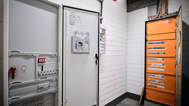 Ein offener Schaltschrank mit Kabeln steht in einem Technikraum vor einem geschlossenen orangefarbenen Schrank