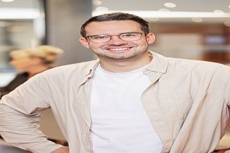 Muž v košili stojí před sedací soupravou a usmívá se do kamery
