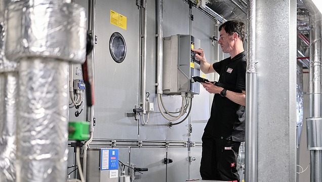 Person steht auf einer Leiter und arbeitet mit Werkzeugen an einem Wand-Bedienfeld in einem industriellen Umfeld mit Rohren, Kabeln und Maschinenkomponenten