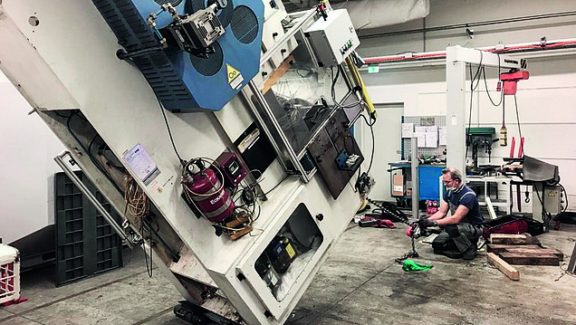 Ein Techniker arbeitet in einer Werkstatt an einem schräg stehenden Maschinenblock, umgeben von Werkzeugen und technischer Ausrüstung.