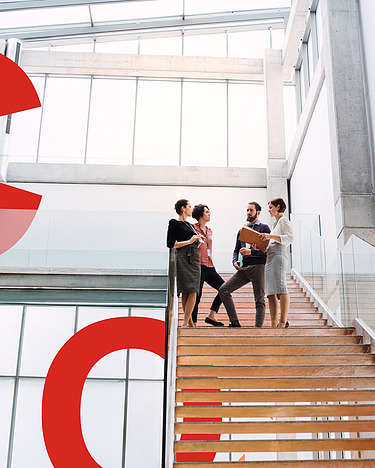 Image of several people from Sustainability Consulting standing on a wooden staircase in a tall building. Next to them are large red signs spelling out C, O and 2.