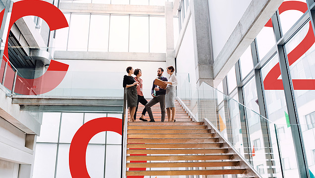 Image of several people from Sustainability Consulting standing on a wooden staircase in a tall building. Next to them are large red signs spelling out C, O and 2.