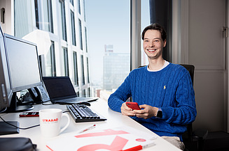 Männliche Person sitzt lächelnd an einem Schreibtisch in einem modernen Büro und hält ein rotes Smartphone in der Hand.