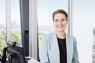 Person in hellblauem Blazer und schwarzem Oberteil steht vor einem Fenster mit unscharfer Stadtlandschaft im Hintergrund