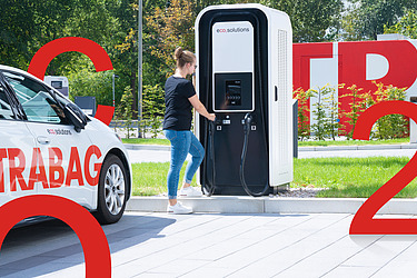 Bild von einem E-Auto das von einer Frau an einer E-Ladesäule geladen wird. Daneben sind große rote Aufsteller von C, O und 2 zu sehen.
