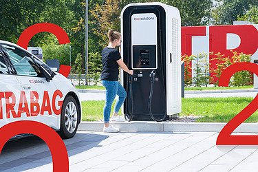 Bild von einem E-Auto das von einer Frau an einer E-Ladesäule geladen wird. Daneben sind große rote Aufsteller von C, O und 2 zu sehen.