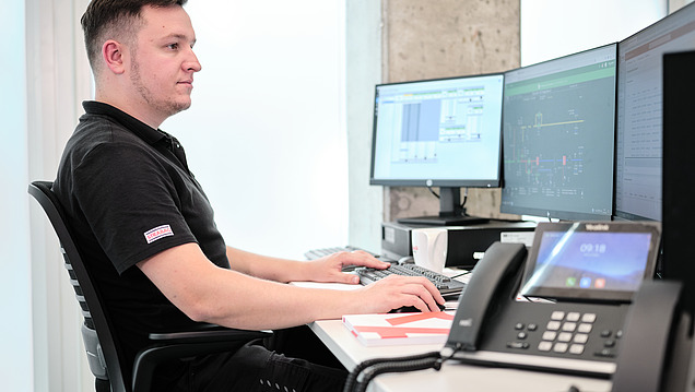 Ein Mann sitzt an einem Schreibtisch und arbeitet an zwei Computermonitoren, während ein Telefon auf dem Tisch steht.