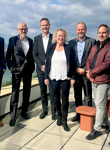 Photo of seven people standing side by side on a roof in the sunshine, smiling at the camera