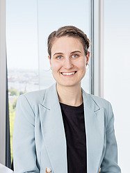 Portrét Mony Gärtnerové - Příjemná žena ve světle modrém saku se usmívá v moderní kanceláři s výhledem na město, pracovní portrét, profesionální.