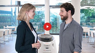 Náhled videa na Yotube: žena a muž stojí před úklidovým robotem v jídelně a stojí proti sobě