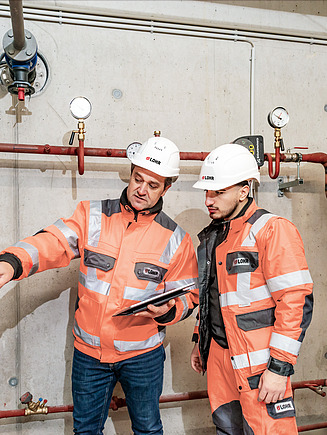 Foto von zwei Personen in Schutzkleidung, die an einer Anlage arbeiten und in ein Tablet eintragen