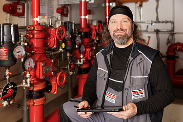 Ein STRABAG-Techniker mit Tablet steht vor roten Rohrleitungen und Ventilen in einem technischen Raum.