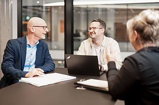 Three colleagues in a meeting, two men talking on laptops, woman from behind, modern office environment.