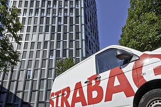 Modernes Hochhaus mit wellenförmiger Fassade, im Vordergrund ein STRABAG-Firmenwagen 