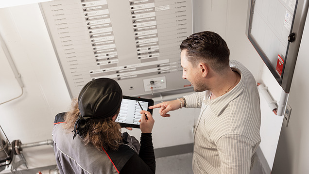 Zwei Personen stehen vor einem großen Schaltschrank und analysieren Daten auf einemTabletut