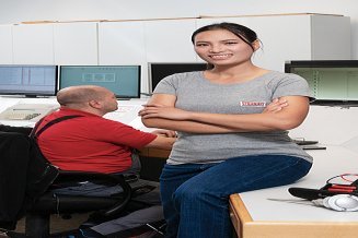 Elektrotechnikerin sitzt mit verschränkten Armen am Tischrand in Gebäudeleittechnik mit zwei Kollegen an Bildschirmen im Hintergrund