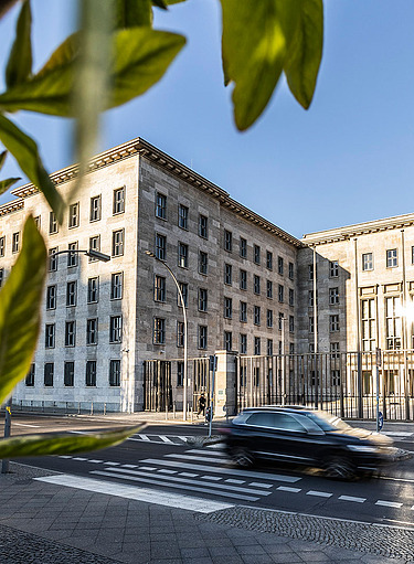 Bild von einem historischen Gebäude, die Vorfahrt ist beflaggt, auf der Straße ist ein vorbeifahrendes Auto zu sehen