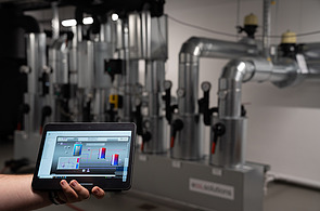 Bild von einem Technikraum und im Vordergrund hält eine Hand ein Tablet